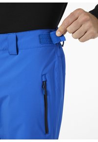 Modré venkovní kalhoty s texturovaným povrchem, nastavitelným páskem na suchý zip a bočním zipovým kapsou, zobrazené v detailním pohledu.