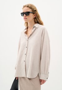 Linen button-up shirt in light beige, featuring a relaxed fit, single chest pocket, and dark buttons. Models paired with ribbed skirt.