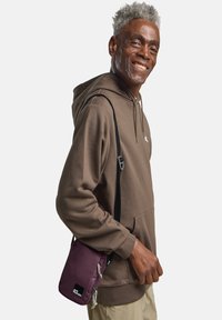 Paarse crossbody tas gemaakt van gladde stof, met een ritssluiting en een zwart logo-label. Model draagt een bruine hoodie en beige broeken.