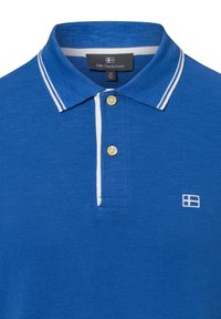 Niebieska koszulka polo z teksturowanej tkaniny, z białym prążkowanym kołnierzykiem, żółtymi guzikami i małym białym logo po lewej stronie.