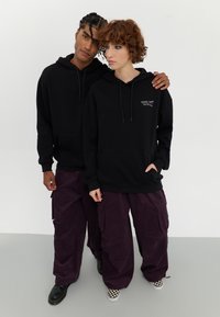 Felpe nere con cappuccio, tasca frontale e cordini, abbinate a pantaloni cargo in corduroy viola scuro e scarpe da ginnastica a scacchi.