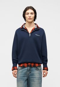 Νεαρός άνδρας φορεί μπλε σκούρο φούτερ με φερμουάρ μέχρι τη μέση πάνω από κόκκινο και μαύρο πουκαμισάκι καρό, συνδυασμένο με ανοιχτό μπλε τζιν, στέκεται μπροστά σε απλό φόντο.