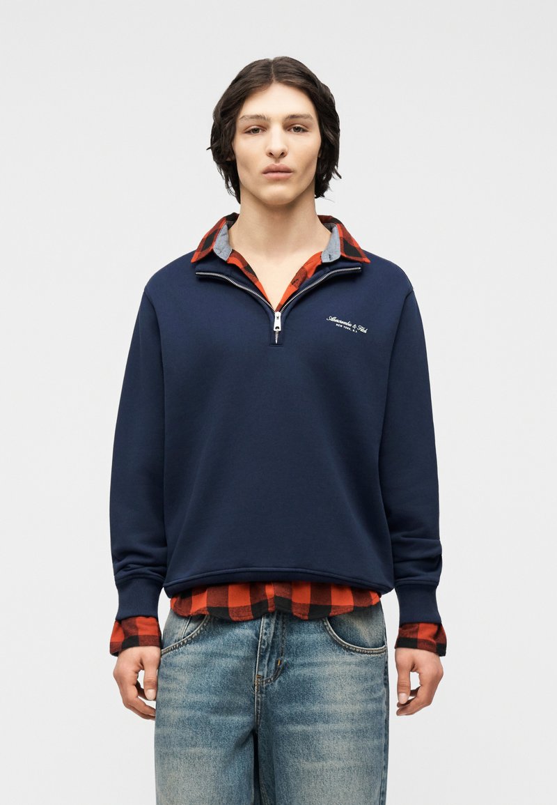 Νεαρός άνδρας φορεί μπλε σκούρο φούτερ με φερμουάρ μέχρι τη μέση πάνω από κόκκινο και μαύρο πουκαμισάκι καρό, συνδυασμένο με ανοιχτό μπλε τζιν, στέκεται μπροστά σε απλό φόντο.