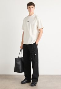 Helles T-Shirt mit kurzen Ärmeln und schwarzem Logo, schwarze Cargo-Hosen mit Seitentaschen, schwarze Ledertasche und schwarze Sneakers.