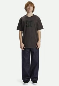 Camiseta gráfica preta com um grande logótipo, combinada com jeans de perna larga azul-escuro e ténis pretos, de pé contra um fundo simples.