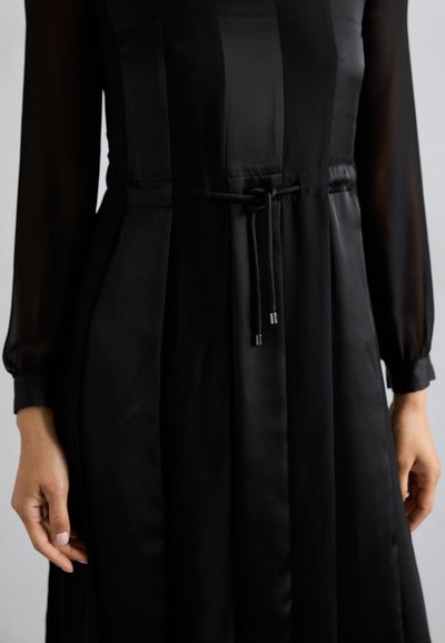 Robe noire en satin avec de longues manches transparentes, dotée d'une taille à cordon, d'une jupe plissée et d'une texture lisse.