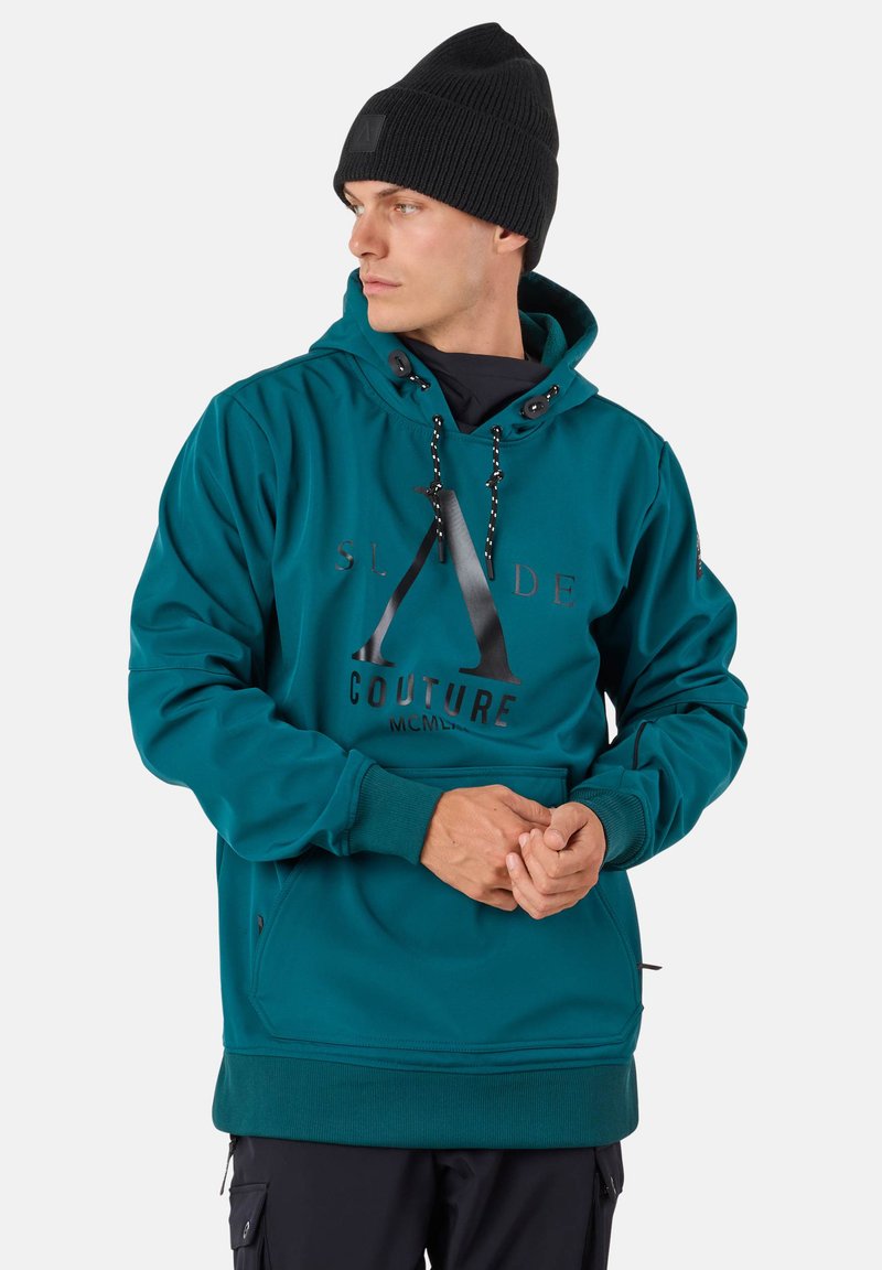 Man draagt een groenblauwe hoodie met zwart logo en zwarte muts, kijkt naar links, handen in elkaar gevouwen, staande voor een witte achtergrond.