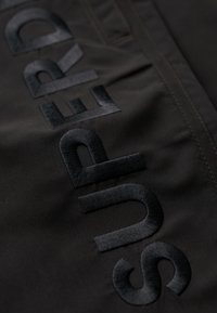 Il tessuto nero presenta la scritta ricamata "SUPERD" in spessori variabili, con una texture liscia. I punti di cucitura sono scuri, completando l'insieme del design.