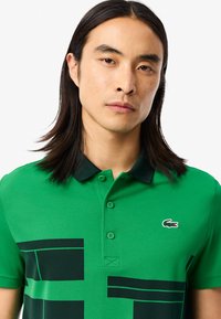 Grünes Poloshirt mit schwarzem Kragen und Knopfverschluss, ausgestattet mit einem großen geometrischen schwarzen Muster und einem kleinen gestickten Logo auf der Brust.