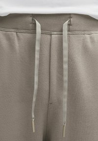 Close-up van de tailleband van beige joggingbroek met ribbel-elastiek en platte trekkoorden die aan de voorkant hangen.