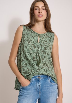 Femme portant un chemisier vert sans manches à motif cachemire avec des liens à pompons et un jean bleu clair, debout avec une main dans la poche.