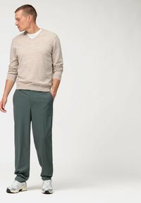Maglione beige a maglia con scollo a V, abbinato a pantaloni verde scuro. Il modello è in piedi con delle sneaker grigie, mani in tasca, e guarda di lato.