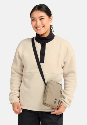 Jonge vrouw die een beige fleece met zwarte kraag en zwarte broek draagt, glimlacht en een beige schoudertas met logo vasthoudt.