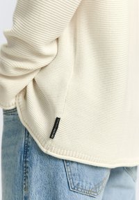 Close-up van een persoon met een crèmekleurige gebreide trui met een zwart etiket waarop "DENIM Tom Tailor" staat en lichtblauwe denim jeans.