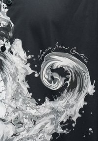 Μαύρο ύφασμα με λευκό και γκρι σχέδιο πιτσιλιάς που μοιάζει με κύμα νερού, με καλλιγραφικό κείμενο "Versace Jeans Couture."