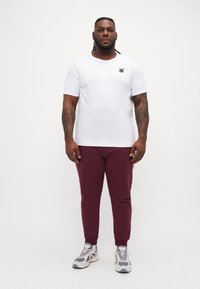 Hvit kortermet skjorte med rund hals og en liten svart logo, kombinert med bordeaux slim-fit bukser og grå treningssko.