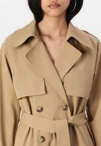 Beige trenchcoat met brede revers, dubbele knopen, een ceintuur in de taille en een gladde textuur. Opmerkelijke details zijn grote knopen en riemlusjes.