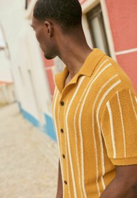 Camisa de manga corta, con cuello, en color mostaza con rayas blancas, tejido texturizado y botones negros. Modelo mostrado de lado.