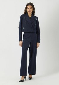 Granatowy garnitur w paski z krótką podwójną marynarką ozdobioną kwiatowymi detalami oraz szerokimi spodniami. Czarne szpilki.