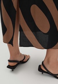 Braun und schwarz gemustertes Kleid mit einem seitlichen Schlitz, kombiniert mit schwarzen, geschnürten Sandalen mit quadratischer Sohle und minimalistischer Gestaltung.