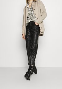 Beige wollenmixjas met bruine knopen, bloemenprint-shirt en zwarte leren broek, gecombineerd met stijlvolle zwarte enkellaarsjes.