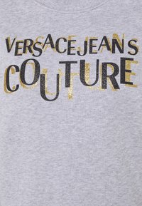 Versace Jeans Couture logo in overlapping black and gold glitter letters on light gray fabric background.