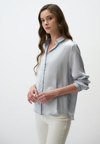 Hellbluse Satinbluse mit durchgehender Knopfleiste, weichem Kragen und hochgeschobenen Ärmeln. Sanfte Textur und entspannter Schnitt.