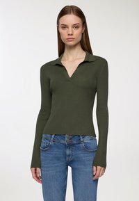 Oliwkowy żeberkowy top z dekoltem w serek i kołnierzykiem. Długie rękawy i dopasowany krój. Noszony z niebieskimi dżinsami z przednimi kieszeniami.