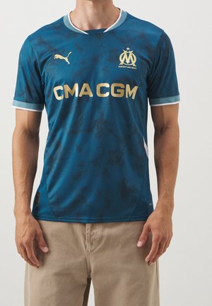 Man draagt een blauw Olympique de Marseille voetbalshirt met goudkleurige Puma-logo en CMA CGM-sponsor, gecombineerd met beige broek.