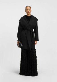 Black wrap coat with a wide collar, belt at the waist, paired with a textured floor-length black skirt featuring layered fringes.