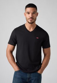 Fekete, rövid ujjú, V-nyakú pamut póló, amelyen a bal mellrészen egy kis piros Levi's logó található. Kék farmernadrággal párosítva.