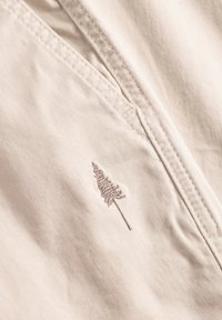 Hellbeige Stoff mit einem gestickten Baumdesign. Sichtbare Nähte und eine glatte Textur, die die detaillierte Stickerei betont.