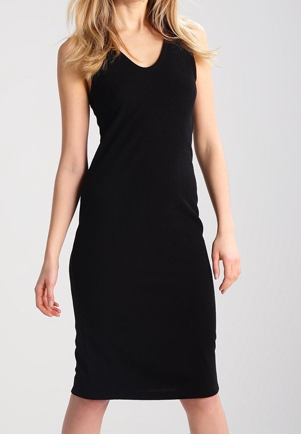 Femme portant une robe noire sans manches longueur genou avec un col en V, debout devant un fond gris clair uni.