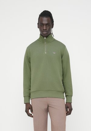 Green sweatshirt with a high collar and half-zip closure. Features ribbed cuffs and hem. Paired with light-colored trousers.