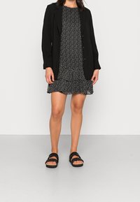 Schwarzer Blazer über einem schwarz-weiß gepunkteten Kleid mit gerafftem Saum. Schwarze Sandalen mit Riemen. Neutraler Hintergrund.