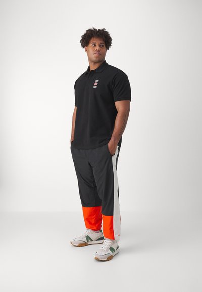 Polo noir avec un petit logo brodé. Pantalons de survêtement noirs amples avec des panneaux latéraux orange, blancs et gris. Baskets blanches avec des accents verts.