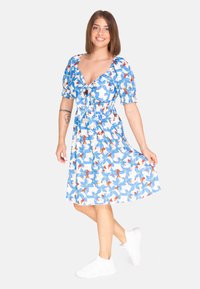 Vestido floral con flores azules y blancas, detalles en marrón, mangas cortas abullonadas, escote profundo y cintura elástica, combinado con zapatillas blancas.