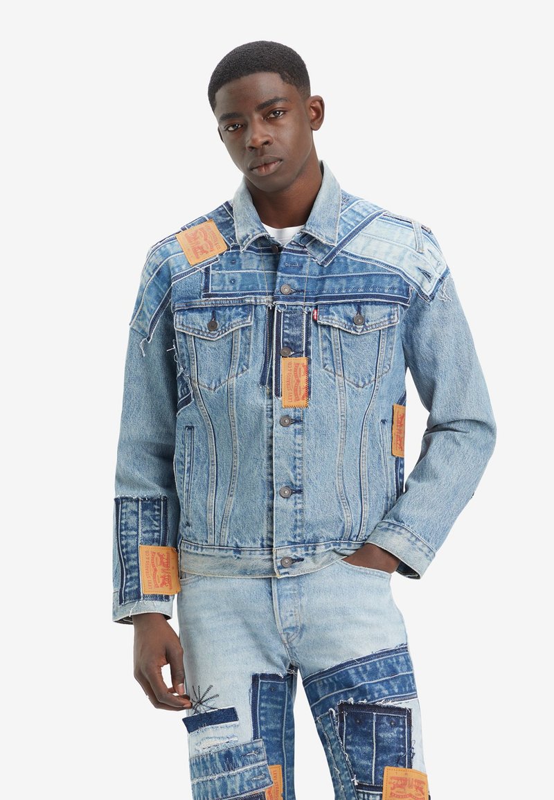 Denim jasje met verschillende tinten blauw, versleten textuur en gele stoffen labels. Klassieke snit met dubbele borstzakken en knopen.