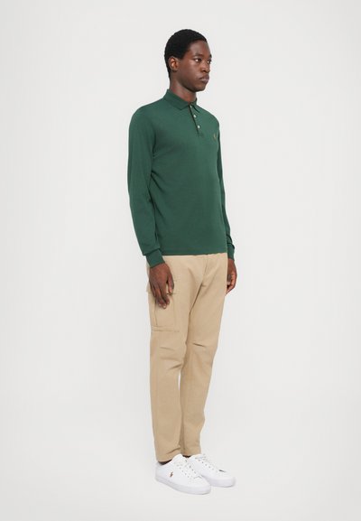 Polo de manga larga de color verde oscuro con cuello y tapeta de botones, combinado con pantalones cargo beige y zapatillas blancas. Diseño simple y práctico.