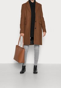 Hnědý kabát z vlněné směsi s klopovým límcem, černý rolák, šedé skinny džíny, černé kotníkové boty a béžová kožená taška.
