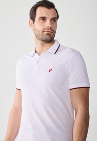 Hvid polo t-shirt med korte ærmer, der har et rødt broderet logo på brystet og røde detaljer på kraven og ærmekanterne.