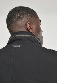 Zwarte jas met een hoge kraag, zilveren rits en een rechthoekige hardware accenten op de achterkant, met een gladde textuur.
