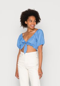 Blusa azul curta com mangas bufantes, laço na frente e barra com babados. Usada com jeans brancos de cintura alta para contraste.
