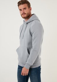 Sudadera con capucha gris, con bolsillo frontal, cordones, puños y dobladillo acanalados. Textura suave, diseño casual, combinada con jeans azules.
