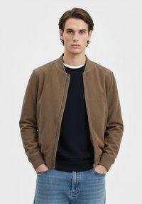 Giacca bomber marrone con colletto arrotondato, chiusura frontale con zip e polsini a costine. Indossata sopra un maglione blu navy e abbinata a jeans azzurri.