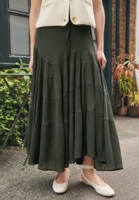 Woman wearing a flowing dark green tiered skirt, cream sleeveless top, and cream ballet flats with ankle straps, standing outdoors on pavement.