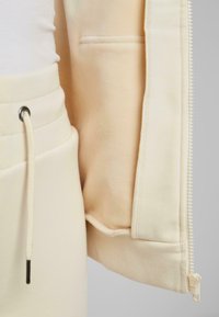 Heledbeez zip-up kapuuts, millel on soonikkoesed ja sile tekstuur, küljetasku ning nööriga detailid taljes.
