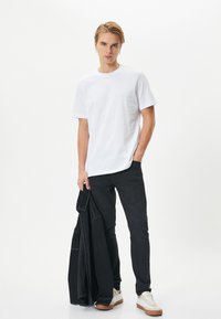 Valkoinen puuvillapaita pyöreällä kaula-aukolle, yhdistettynä mustiin farkkuihin ja vaaleanvärisiin lenkkareihin. Malli pitää mustaa takkia.