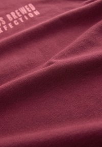 Burgundijas kokvilnas audums ar gludu tekstūru, kurā redzama reljefa balta rakstība "Izpildīta perfektā." Mīksts kritums redzams.