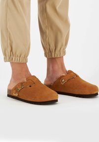 Zoccoli in suede marrone con punta rotonda, fibbia laterale e suola texturizzata, abbinati a pantaloni beige elasticizzati.
